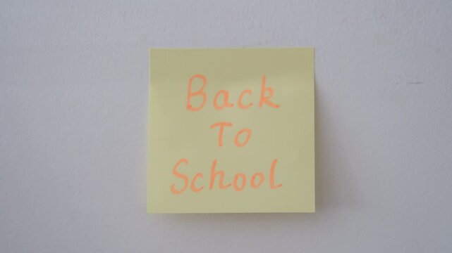 A person's hand pressing a square yellow sticky note onto a plain white wall. The note features the words "Back To School" written in orange handwritten script.
