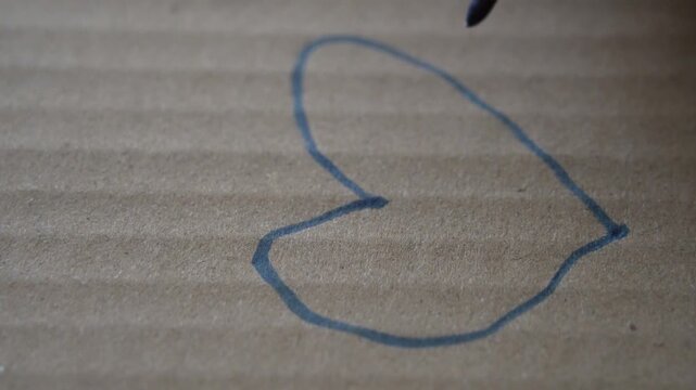 Hand drawing a heart with a blue highlighter on cardboard, representing love, warmth, or simple emotional expression, close-up