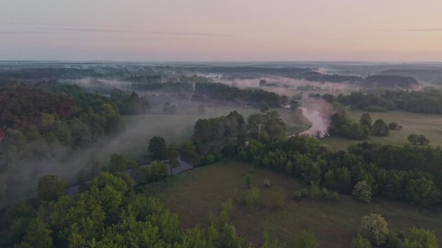 Wide aerial drone view of Wkra river meandering through lowland landscape blanketed in dense morning fog, lush green trees and meadows emerging from mist, soft pastel pink dawn sky on horizon