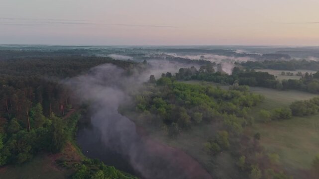 Wide aerial drone view of Wkra river valley covered in thick morning fog drifting through lush green forest and meadows, pine trees on left, soft pastel pink dawn sky stretching to flat horizon