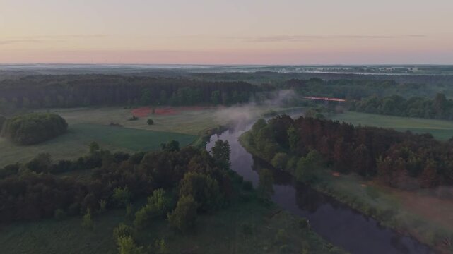 Wide aerial drone view of Wkra river winding through misty lowland landscape with morning mist rising above water, railway bridge visible in background, agricultural fields, forest treeline, dawn sky