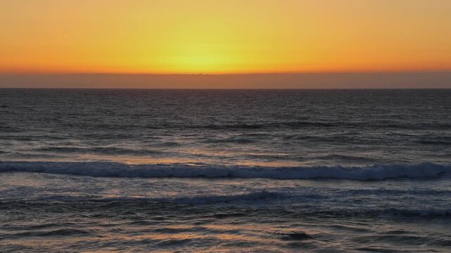 Wide view of vivid orange and golden sunset with sun touching horizon over dark Atlantic Ocean, rolling waves in foreground, Imsouane, Morocco