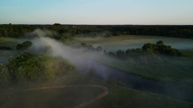 Aerial drone view of Wkra river winding through green meadows with dense morning mist rising above water, dirt path and tree clusters in foreground, forest treeline on horizon, sunrise light