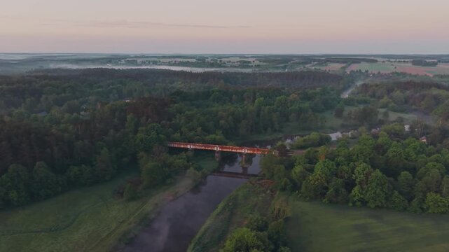 Aerial drone view of rusty railway bridge spanning Wkra river surrounded by lush green forest, morning mist drifting across distant lowland plain, soft pastel pink dawn sky on horizon
