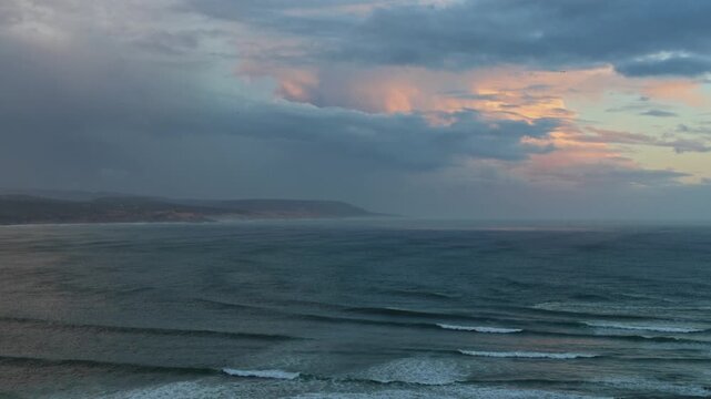 Wide view of dramatic stormy sky with rain shower and pink-lit clouds over dark Atlantic Ocean, rolling waves and rocky coastal headland visible in background, dusk light, Imsouane, Morocco
