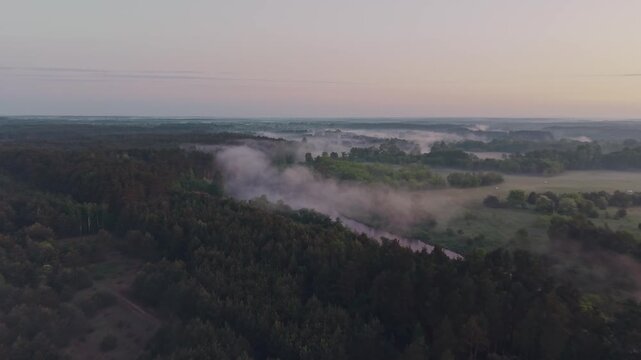 Wide aerial drone view of Wkra river winding through dense forest with morning mist rising above water and treetops, flat lowland plain and soft pastel dawn sky stretching to horizon