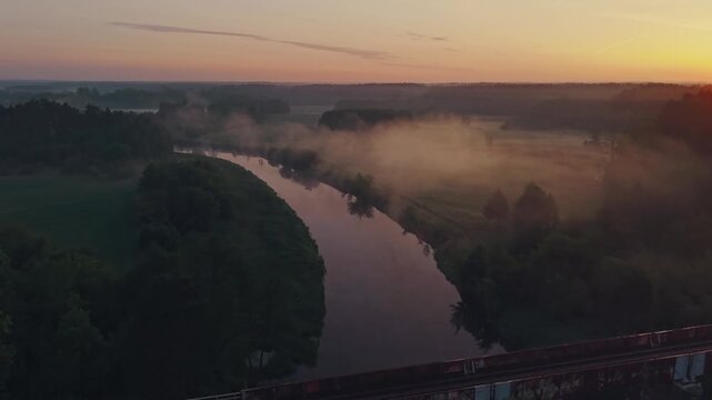 Aerial drone view of Wkra river winding through misty lowland landscape, dense morning fog drifting over water and meadows, vivid orange sunrise sky reflecting on calm river surface
