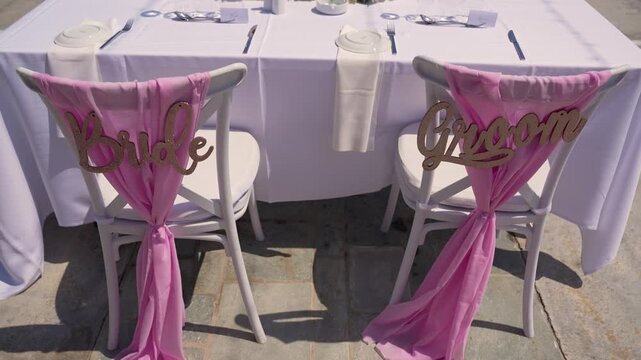 Chairs for the bride and groom sit at a table set for a wedding celebration. The decorations include pink sashes and wooden signs on the chairs.