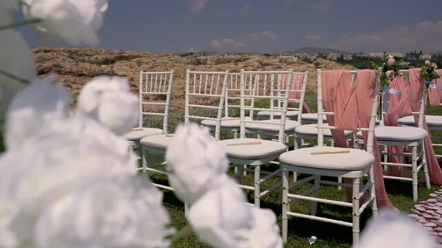 Chairs are arranged for a wedding ceremony on a grassy area. The venue is outdoors, with decorative elements like flowers and fabric draping visible. Sunny weather is present.