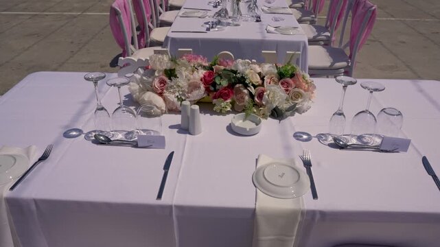 A long table is prepared for a wedding reception. Flowers are arranged in the center. Plates, glasses, and utensils are neatly placed for guests. The location has a simple, open area.