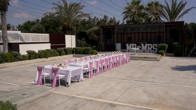 A garden is set up for a wedding with long tables arranged in a row. Chairs are decorated with pink bows. The sun shines on the scene with palm trees in the background.