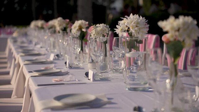 A long table is arranged for an outdoor event. Glasses and plates are placed neatly, and flowers are in vases. The setup is ready for guests in a pleasant setting.