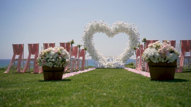 A wedding venue is set up on a green lawn near the sea. White flowers create a heart shape at the front. Chairs are arranged for guests with flower pots on the sides.