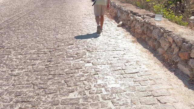 Man walking on a cobblestone path beside an old stone wall wearing shorts and gray sneakers, casual travel stroll, exploration on foot, summer vacation lifestyle.