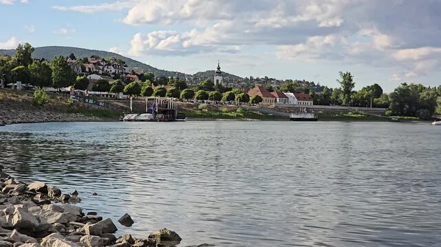 Beautiful static view of the historic town of Szentendre, Hungary, seen from across the Danube River. The picturesque church tower, waterfront, green hills, and dramatic summer clouds create a peacefu