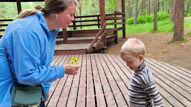 A boy hands a bouquet of wildflowers to his mother on a wooden platform in a forest park. A warm family moment, caring and generational connection.

