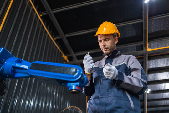 Male engineer wearing yellow helmet inspecting blue robot arm for maintenance in dark industrial factory setting with serious expression and complete concentration.