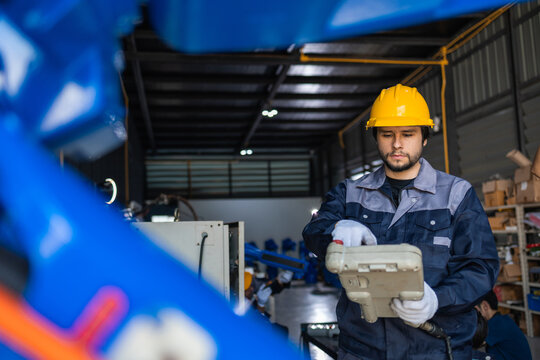 Focused adult Caucasian worker controls industrial automation equipment using manual device for operating mechanical blue robot arm inside modern manufacturing factory