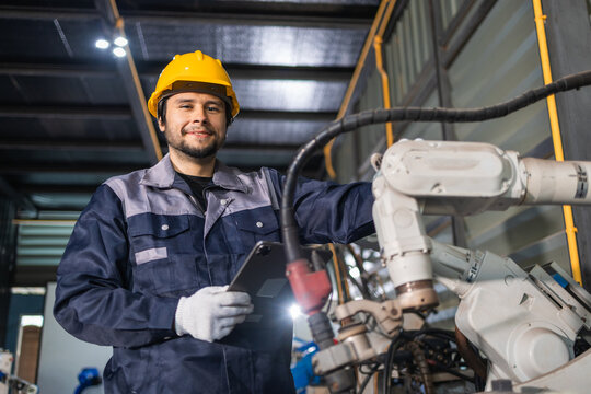 Confident smiling male engineer wearing yellow helmet holding tablet standing by industrial robot arm for modern manufacturing process inside large dark factory building with bright lighting