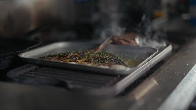 Chef bastes fish on a metal tray in a professional kitchen setting
