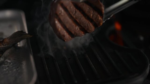 Grilled steak sizzles on barbecue grate with tongs held nearby