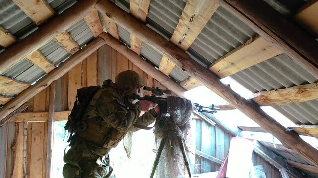 A soldier in camouflage walking up to a camouflaged sniper rifle mounted on a tripod and starting to aim at the enemy.