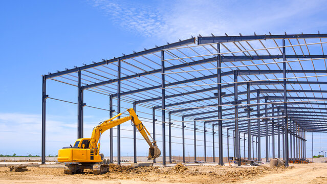 Excavator is working to leveling the ground with industrial building framework structure in factory construction site against blue sky background