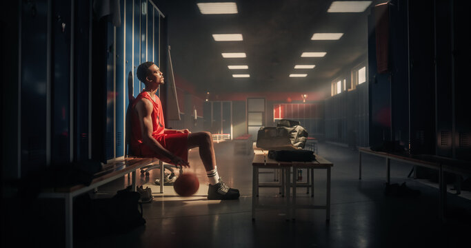 African American Male Basketball Player in a Red Jersey and Shorts Sits Alone on a Wooden Bench, Mentally Preparing Beside a Ball, Duffel Bag and Blue Lockers in a Dark Empty Locker Room