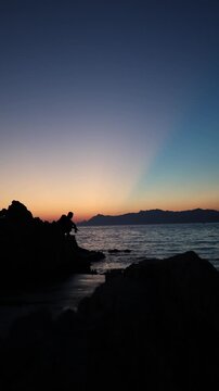 Vertical screen Spinning fishing at sunset follows an angler retrieving lure across calm water as fading daylight creates peaceful evening atmosphere by rocky shore