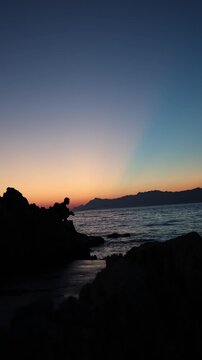 Vertical screen Sunset fishing silhouette on rugged rocks creates peaceful evening mood with calm waves fading light and quiet reflection after a long summer day