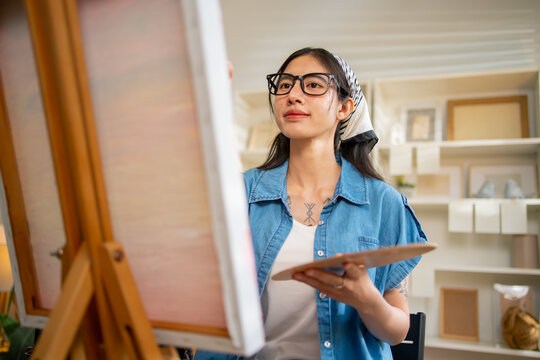 Young Asian woman artist painting on canvas easel with paintbrush in art studio, representing creative professional women designer, fine arts student lifestyle and freelance illustrator
