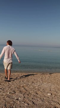 Vertical screen A carefree traveler stretches out his arms while walking beside the calm sea embracing the joy of vacation freedom and a peaceful Mediterranean morning