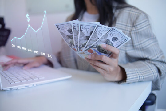 Businesswoman Checking Rising Financial Charts On Laptop While Holding Dollars At Desk