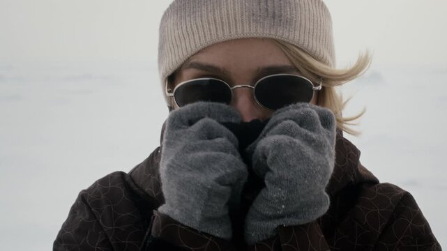 Portrait of Caucasian adult woman in sunglasses and beanie covering face from cold on snowy field. Clip is ideal for winter apparel marketing and outdoor gear commercials