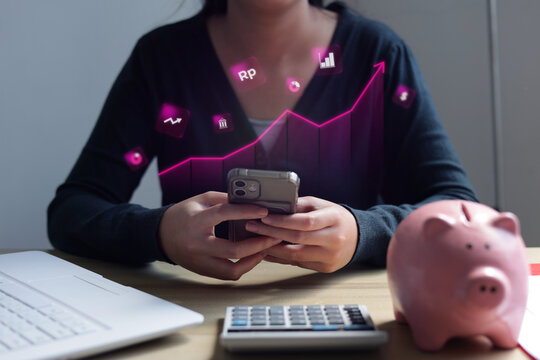 Female Investor Checking Digital Savings On Phone Alongside Laptop Calculator And Piggy Bank