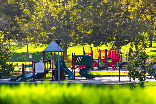 Colorful playground slides and climbing structures stand empty beneath mature trees on a bright green lawn at Carbon Canyon Regional Park.