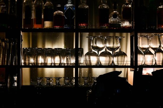 Atmospheric night shot of a cozy bar interior featuring backlit liquor bottles, clean wine glasses, and stemware neatly arranged on dark wooden grid shelves with warm soft lighting.