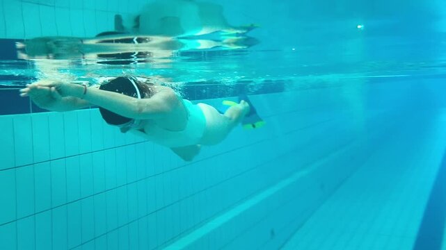 Underwater view of an adult woman practicing freestyle swimming technique in an indoor pool, improving stroke coordination, breathing, confidence, and fitness during training sessions alone