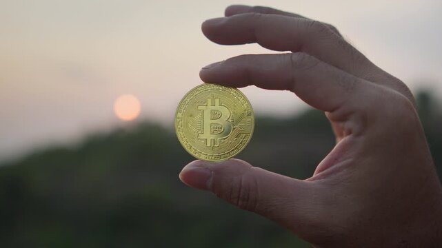 Close up of a hand holding a physical Bitcoin coin against a dramatic sunset sky, symbolizing digital finance, investment, and the future of decentralized currency.