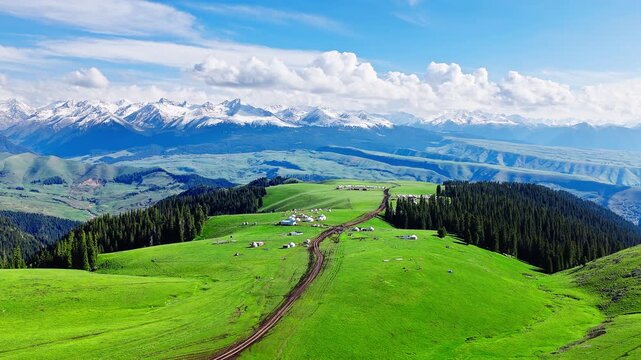 Aerial view of a nomadic yurt camp on rolling green hills with a snow-capped mountain range in the background, Kalajun Grassland, Xinjiang, China.