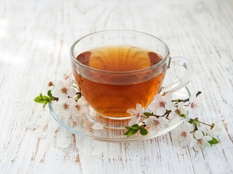 A clear glass cup filled with warm herbal tea decorated with fresh green leaves and small flowers on a rustic wooden table