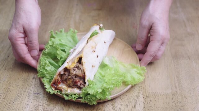 Person holding a wooden plate with a meat shawarma wrap and fresh green lettuce. The savory wrap is filled with grilled meat and red sauce, served on a rustic wooden tabletop background.