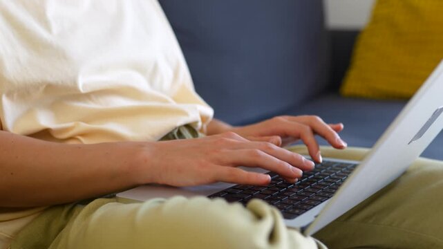 Close-up of woman sitting on a sofa with a laptop, typing on a keyboard. Female hands working on a laptop in a cozy room. Concept of remote work, technology, typing.