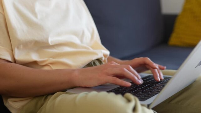Close-up of woman sitting on a sofa with a laptop, typing on a keyboard. Female hands working on a laptop in a cozy room. Concept of remote work, technology, typing.