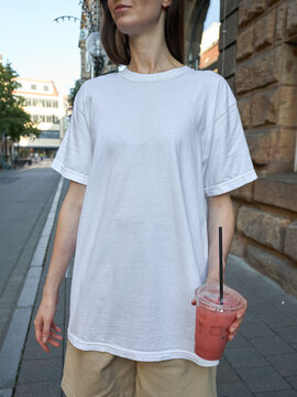 Woman in white t-shirt and beige shorts holding a pink drink on a city street