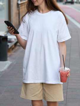 Woman in white t-shirt and shorts holding a phone and a pink drink