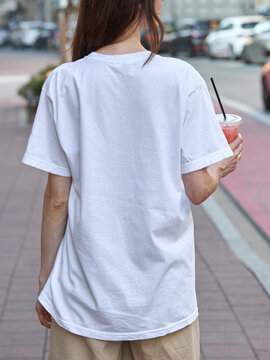 Woman in white t-shirt holding a pink drink while walking on a city street