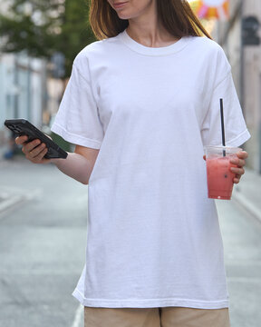 Woman in white t-shirt holding a phone and a pink drink on a city street