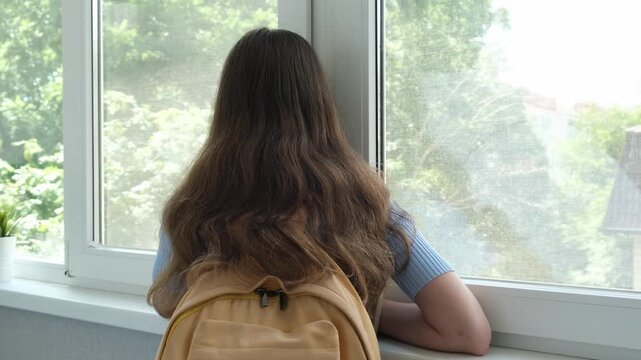 Rear view of thoughtful schoolgirl with backpack looking out of window. Unhappy teen girl alone