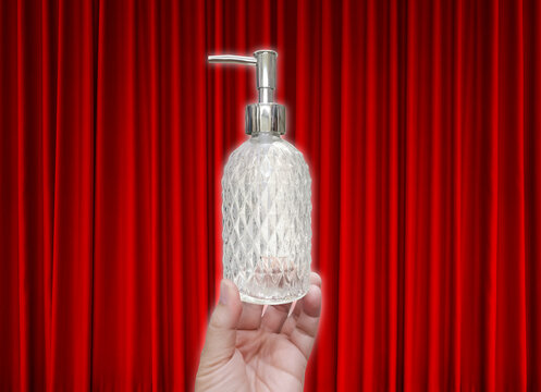 A clear glass perfume bottle with a diamond pattern is held in hand, against a red curtain as a backdrop.